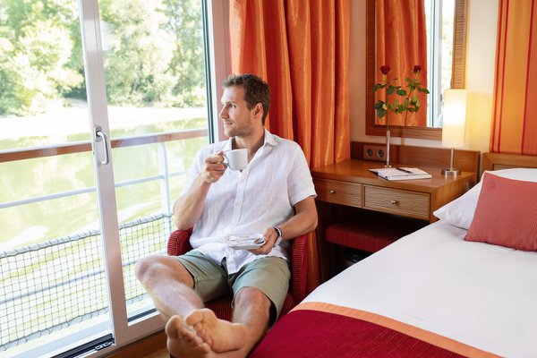 Man in a white shirt and shorts sits on a chair by the window of a cabin and drinks coffee.