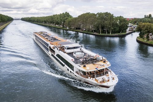 A river cruise ship sails along an urban shore with trees, with a bridge in the background. The sun deck is equipped with a whirlpool, sun loungers, and sun sails.