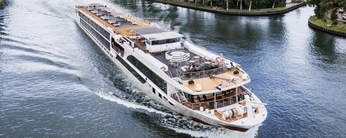 A river cruise ship sails along an urban shore with trees, with a bridge in the background. The sun deck is equipped with a whirlpool, sun loungers, and sun sails.