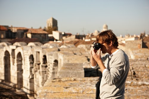 A man stands on a historic wall in Arles and photographs something. In the background, the yellow buildings of the city.