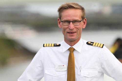 Man in a white shirt with epaulettes and a brown tie with the A-ROSA logo, standing with one hand on a railing and the other on his hip.