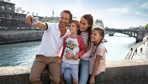 Family with father, mother, son, and daughter takes a selfie on a bridge over the river in Paris in sunny weather.