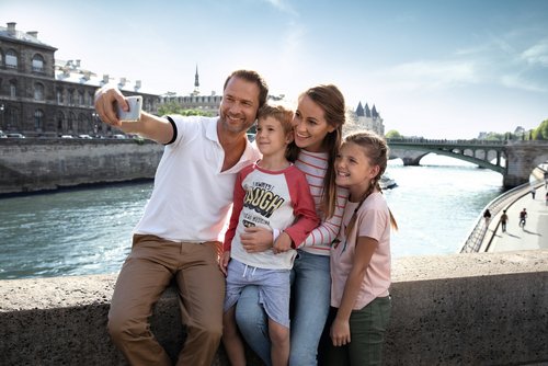 Family with father, mother, son, and daughter takes a selfie on a bridge over the river in Paris in sunny weather.