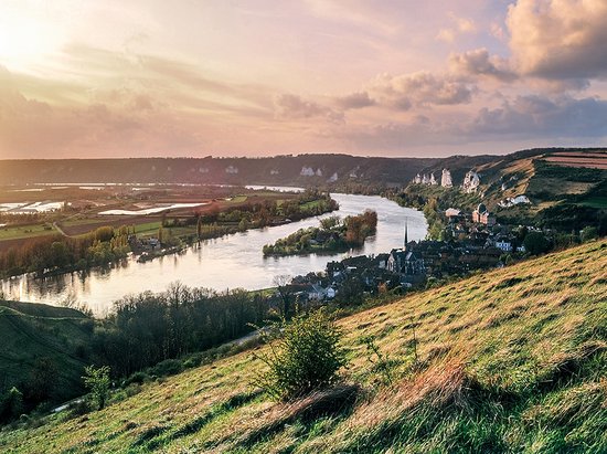 River Seine, winding through green hills and the village of Les Andelys with its church and rocky landscape at sunset.