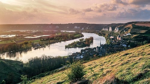 River Seine, winding through green hills and the village of Les Andelys with its church and rocky landscape at sunset.