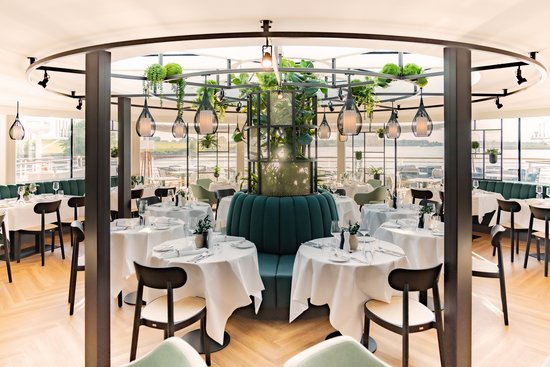 Modern onboard restaurant of the A-ROSA SENA with round tables, white tablecloths, green benches, and hanging plant lamps in a bright room with large windows