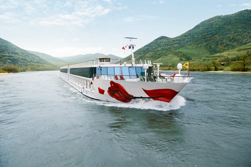 A large river cruise ship of A-ROSA on the Danube, with several decks and red accents, sails past a wooded riverside landscape.