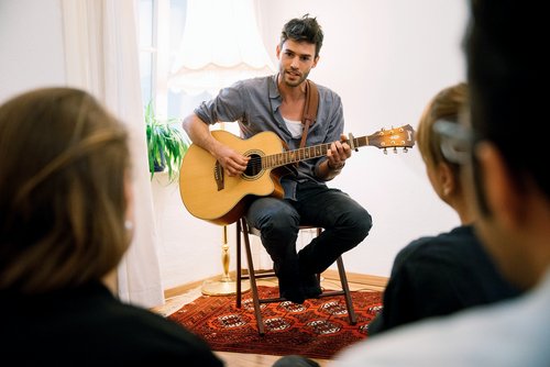 Musician Henrik Wiards plays acoustic guitar at an intimate concert. He sits on a chair facing a small group of listeners in a living room with carpet and plants.