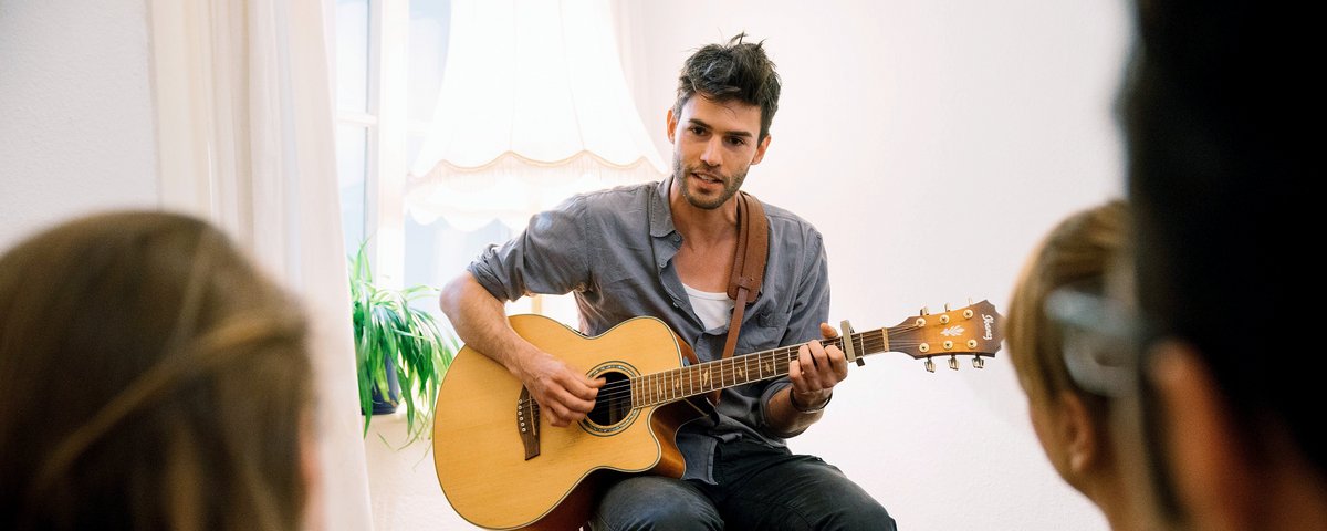 Musician Henrik Wiards plays acoustic guitar at an intimate concert. He sits on a chair facing a small group of listeners in a living room with carpet and plants.