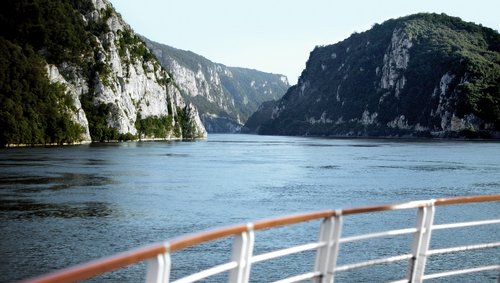 View from a river cruise ship of the vegetated rocky landscape in the Danube Delta. In the foreground is a railing and behind it the quiet river.