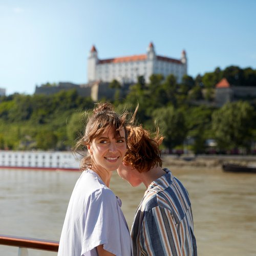 Two women stand at the railing on a river cruise ship on the Danube, looking at another ship and Bratislava Castle on a wooded hill in the background.