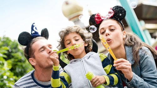 Two children and their father at Disneyland Paris. The father and the girl on the right are wearing Mickey Mouse ears on their heads. The boy in the middle and the girl are blowing soap bubbles.